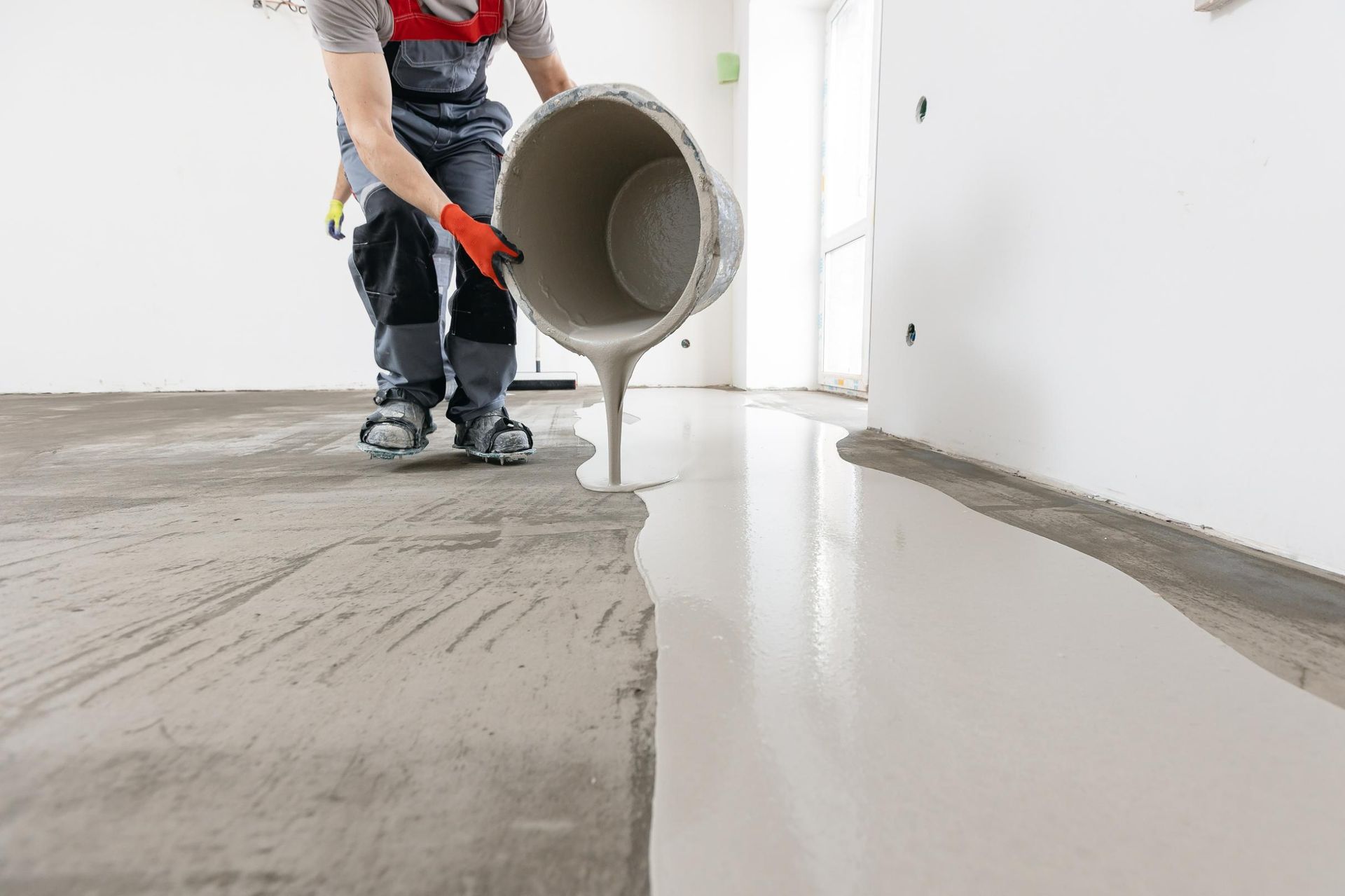 A construction worker pours leveling compound onto a concrete floor, preparing it for a new finish.