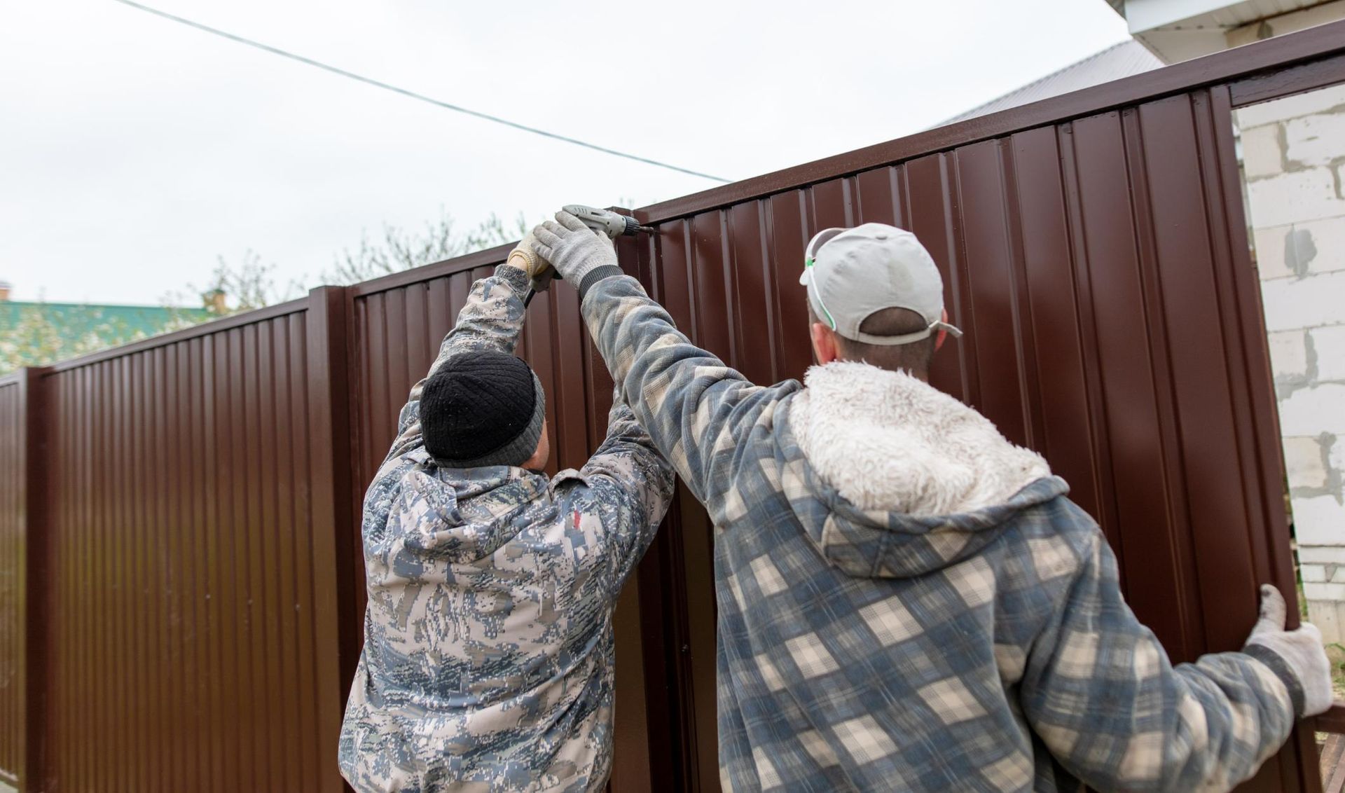 Two workers installing a brown metal fence panel outside a building on a cloudy day.