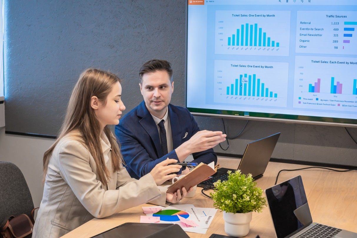 A Man And A Woman Are Sitting At A Table Looking At A Clipboard.