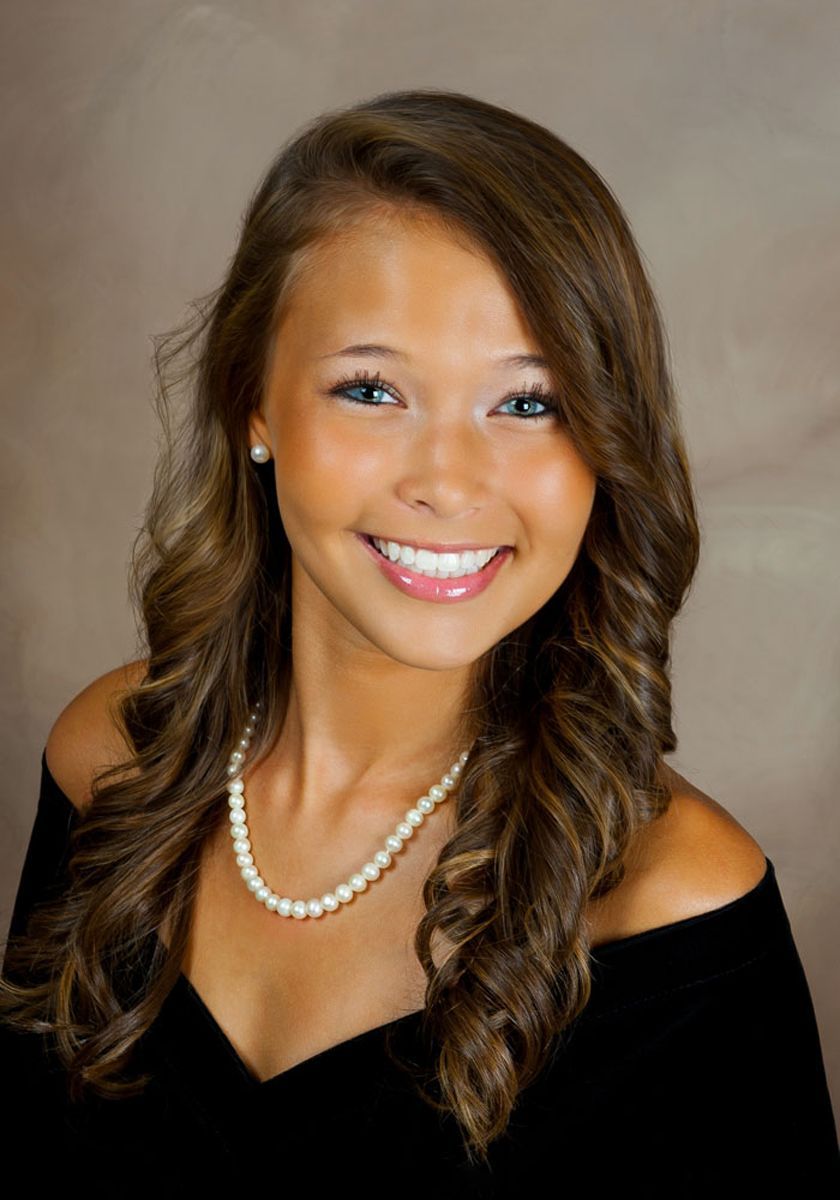 Portrait of a smiling woman with wavy brown hair, wearing a black top and pearl necklace against a neutral background