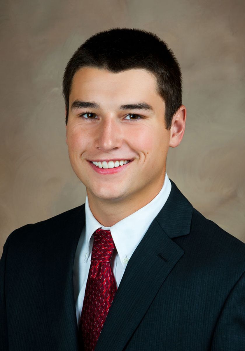Man in a dark suit, white shirt, and red tie, smiling against a neutral studio background