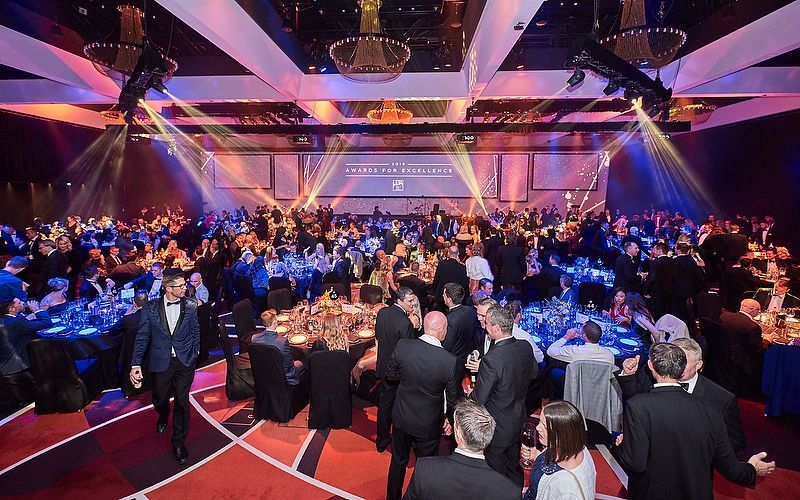 Crowded formal banquet with people seated at round tables under colorful stage lighting in a large hall