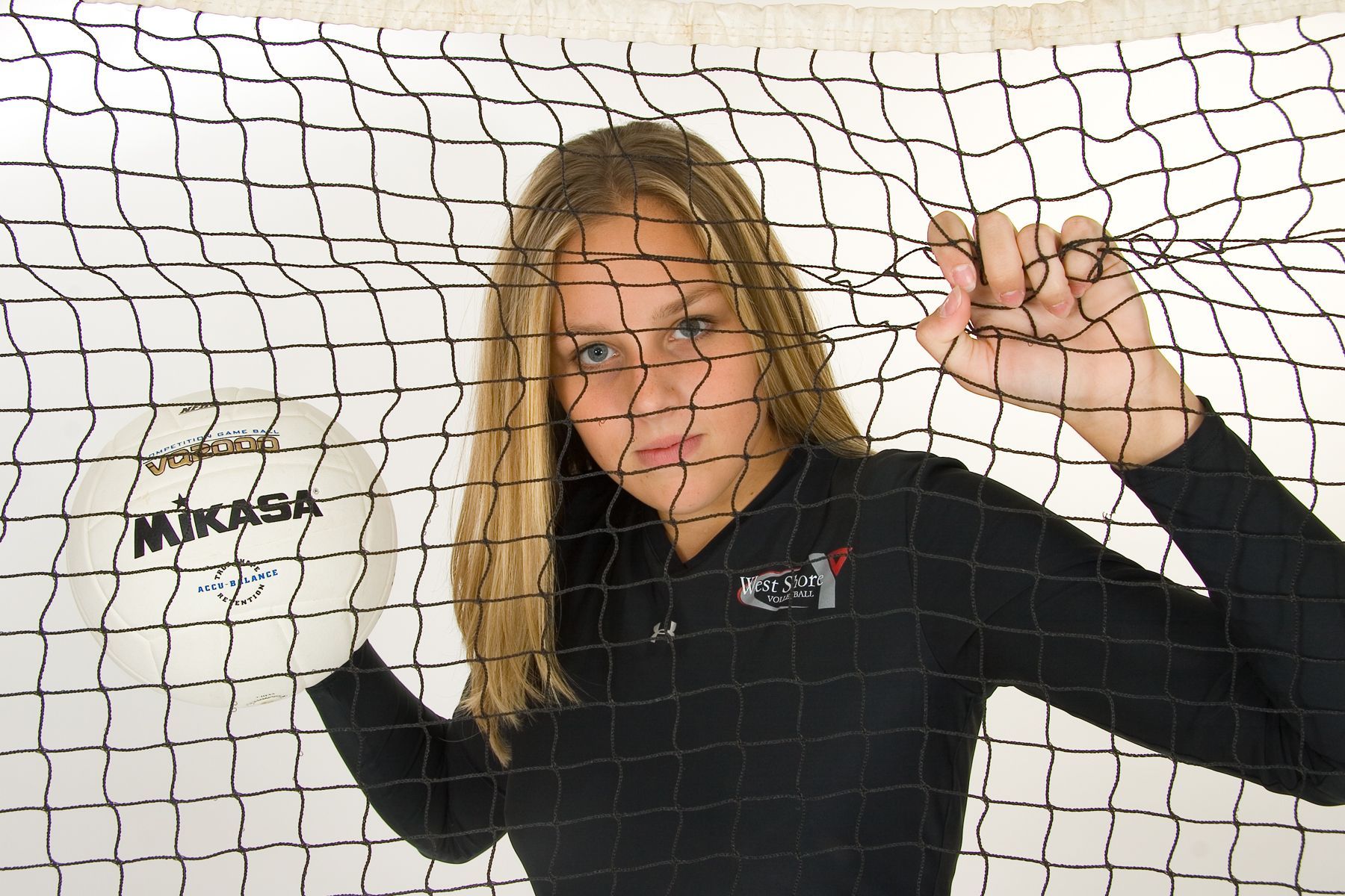 Woman standing behind volleyball net, holding up her hands against a white wall.