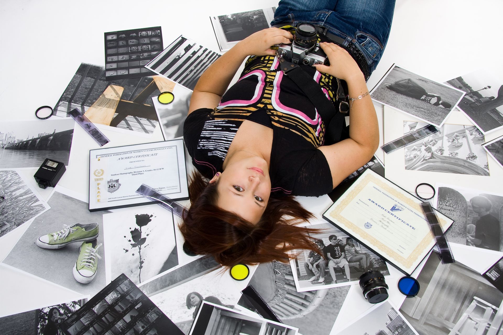 Woman lying on scattered black-and-white photos, holding a camera, with certificate and contact sheet nearby