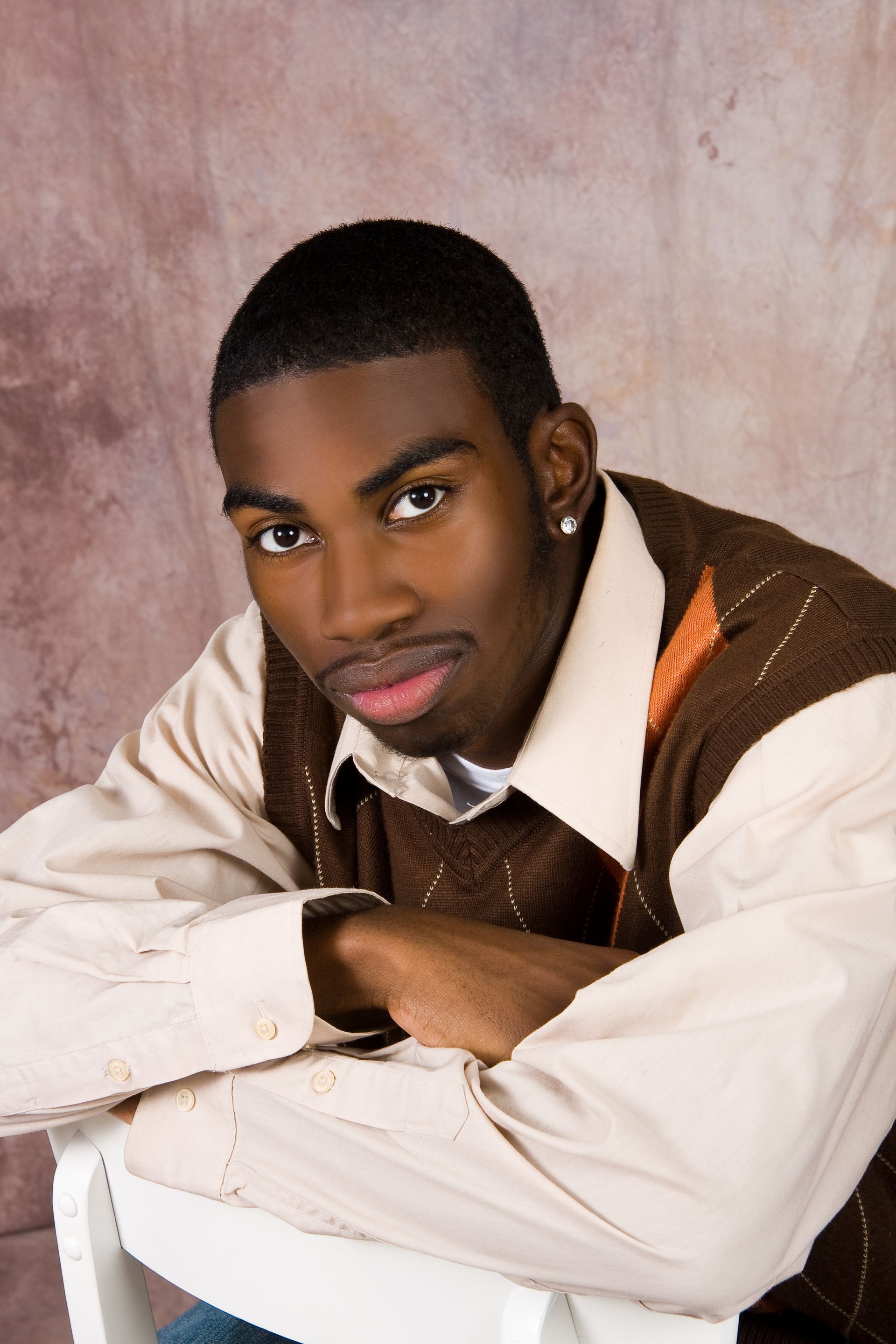 Portrait of a man in a white shirt and brown vest, leaning forward with folded arms against a soft pink backdrop