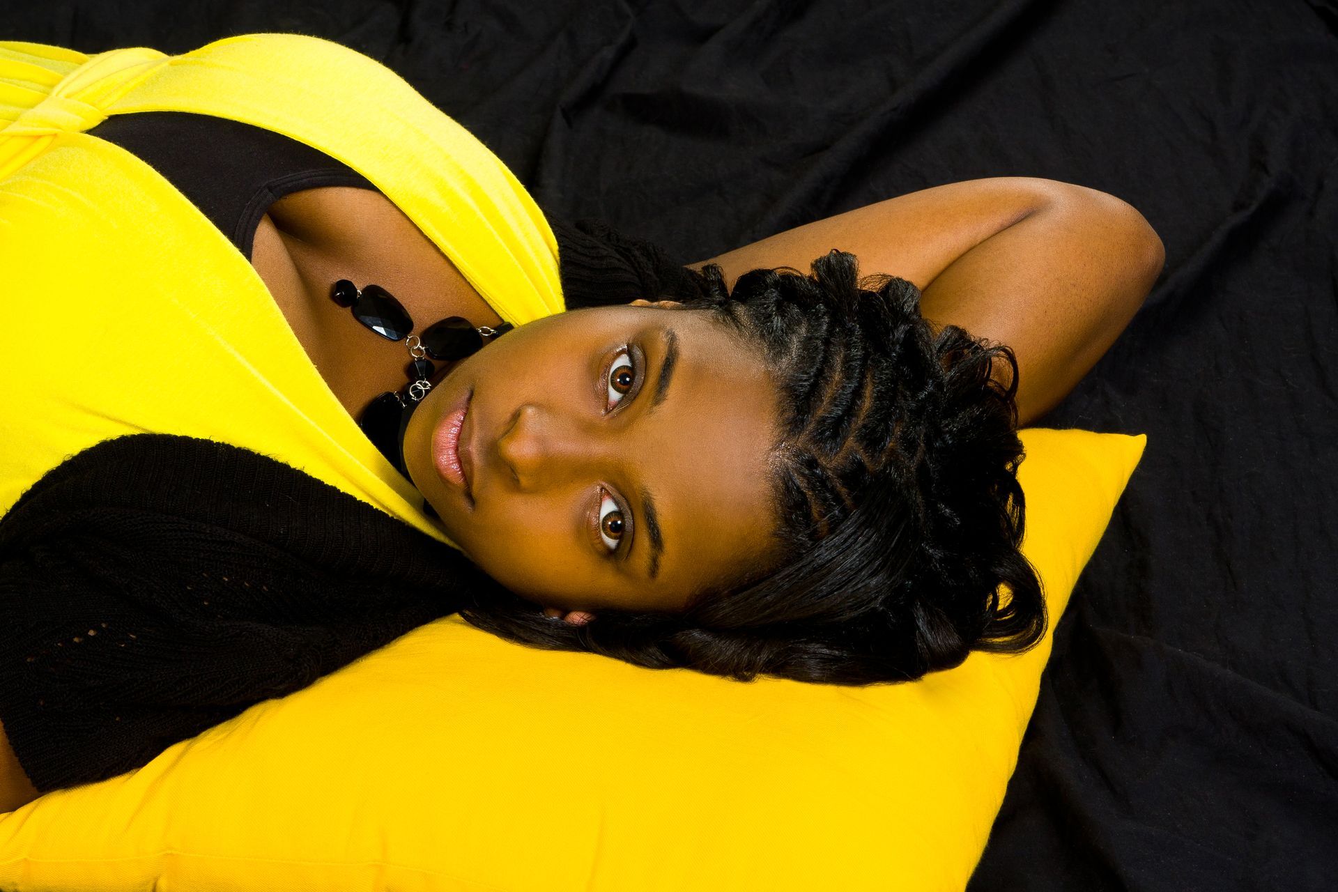 Person lying on a bright yellow pillow against a black background, looking at the camera.