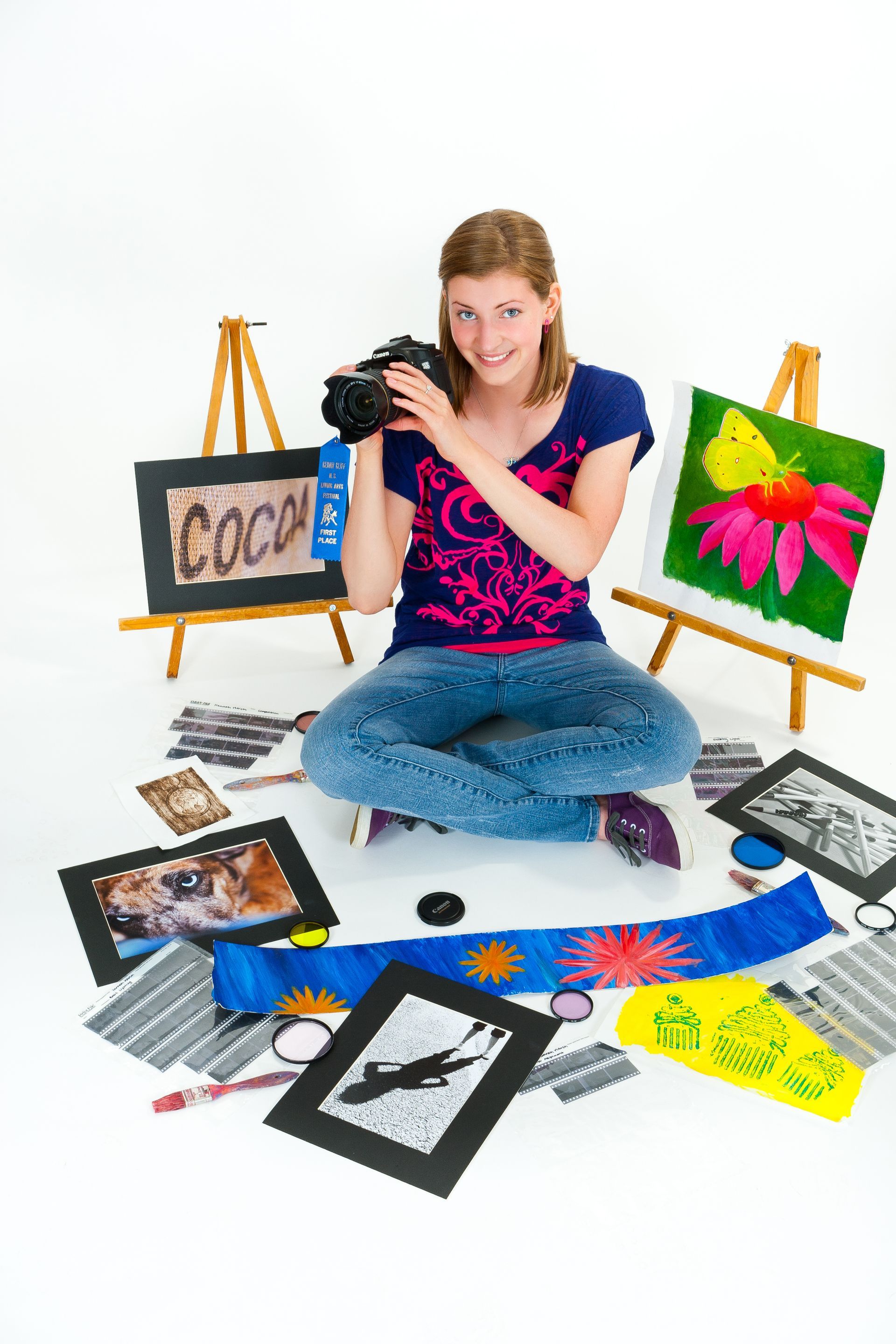 Artist sitting on the floor with paintings and a camera, surrounded by colorful artwork on easels.