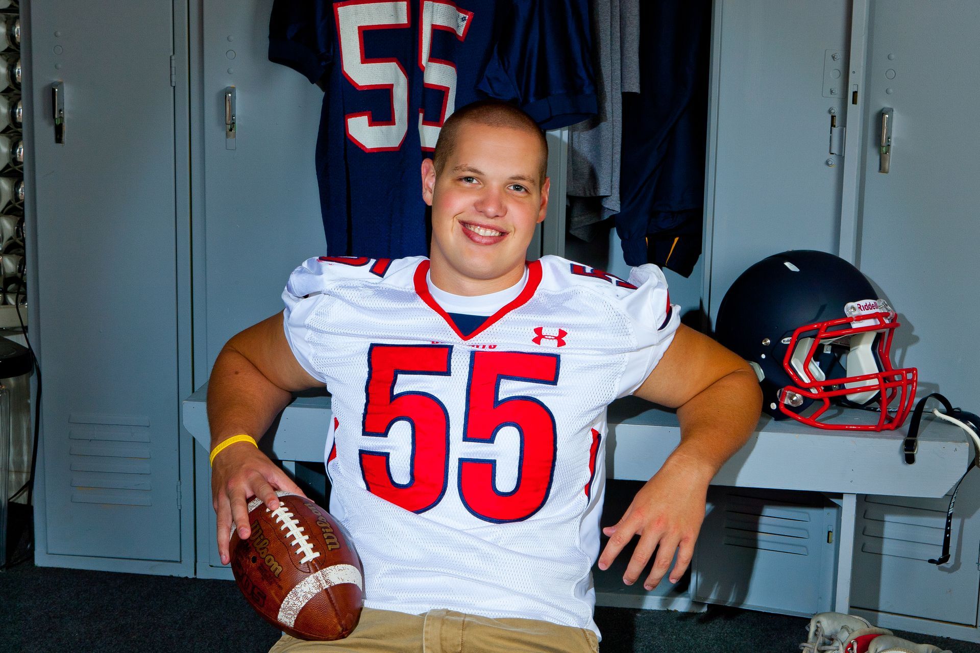 Football player in white jersey number 55 holding a football in a locker room, smiling at camera