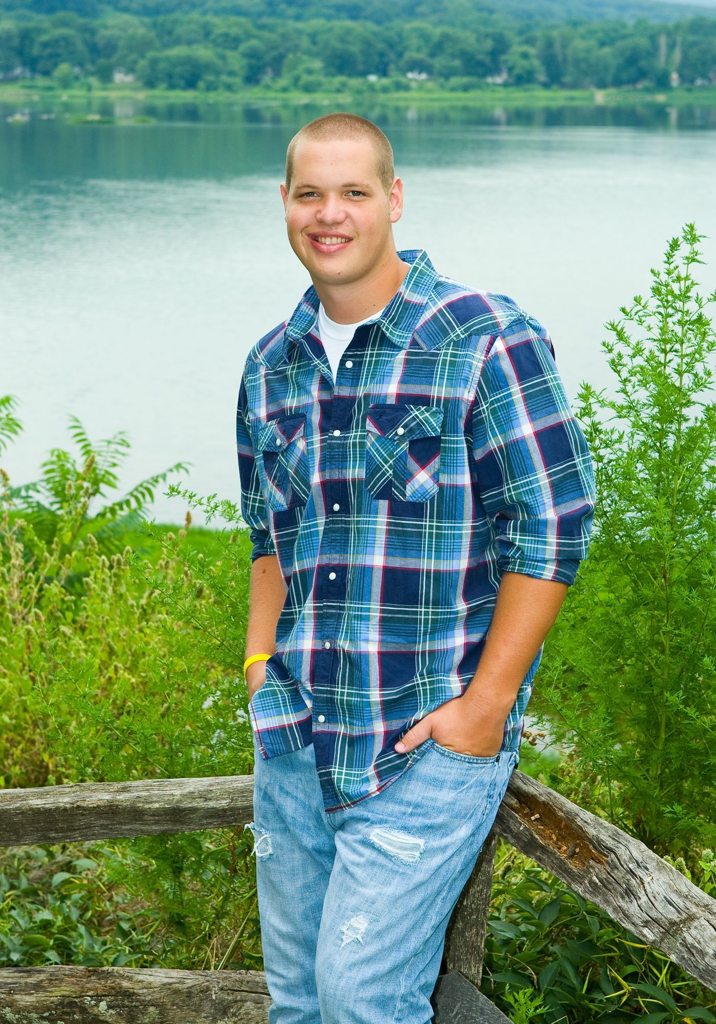 Man in a blue plaid shirt and jeans standing by a lakeside fence with trees in the background