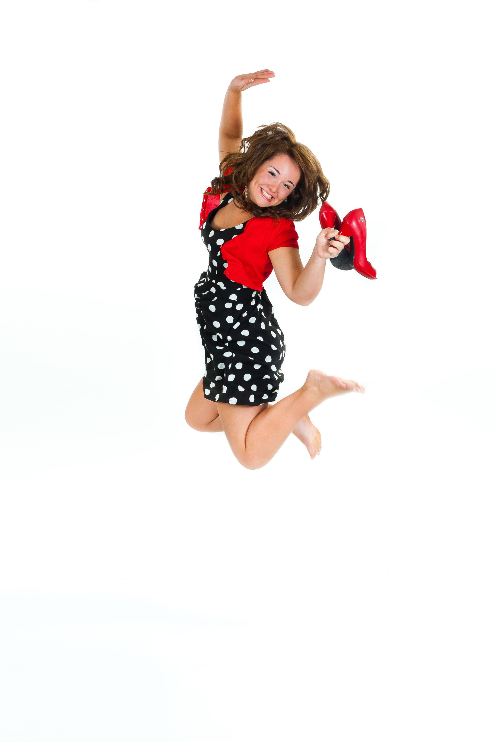 Woman in a black polka-dot dress and red top jumping against a white background, holding red shoes