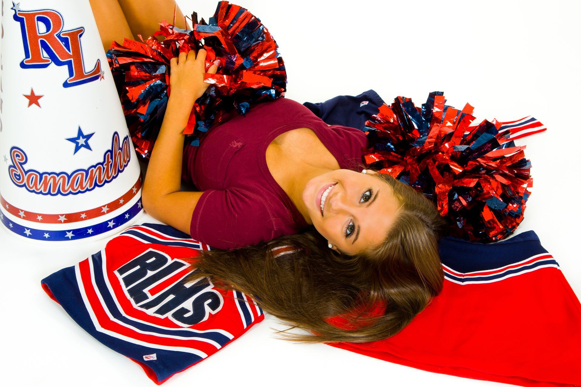Cheerleader posing with red, white, and blue pom-poms beside a red-white-blue mat and decorated megaphone
