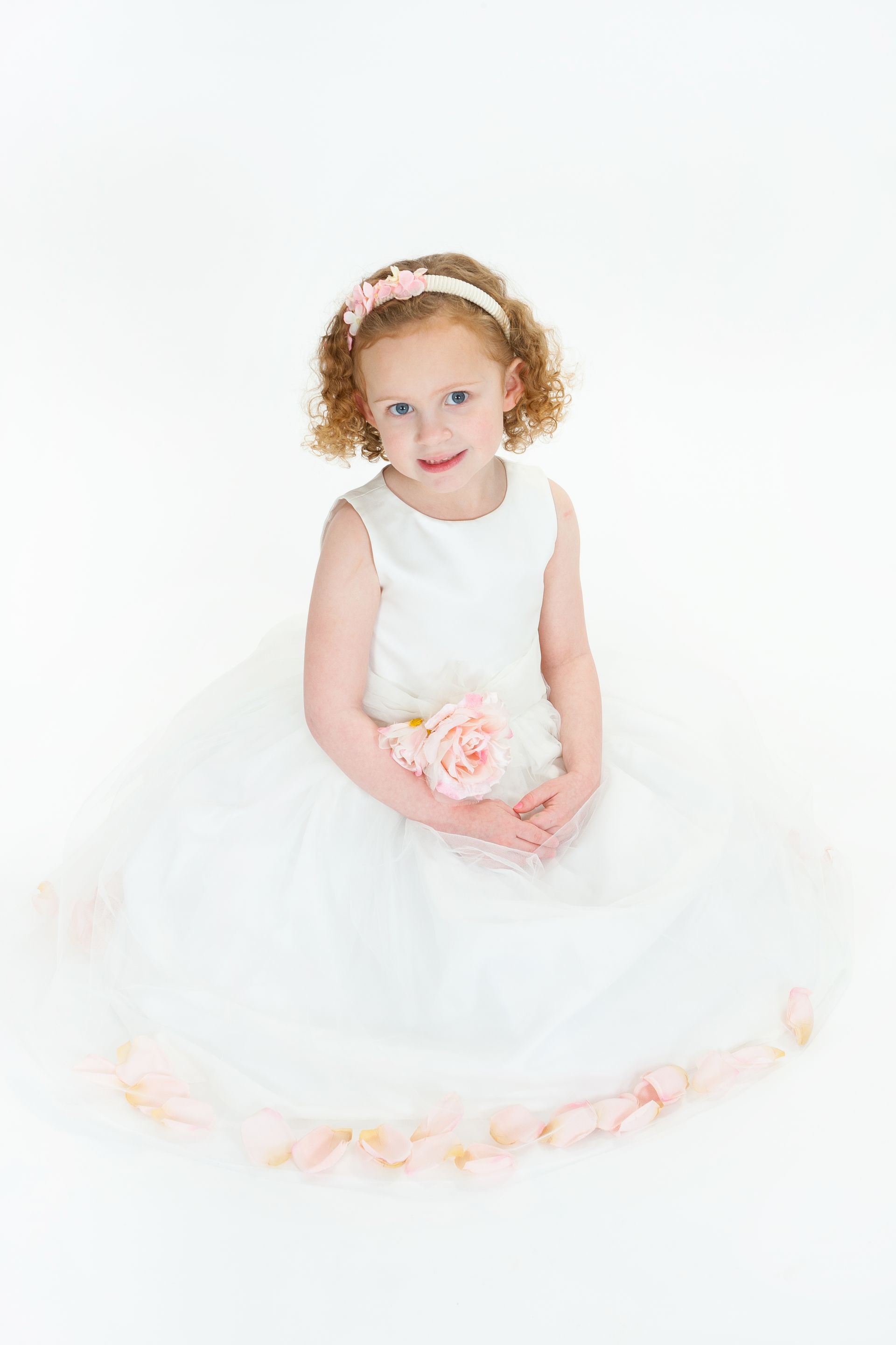 Young child in a white dress holding pink flowers, seated against a bright white background.