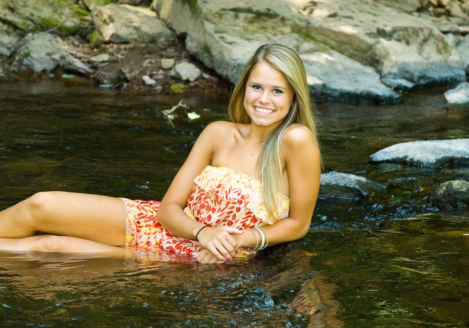 Woman reclining in shallow stream, smiling in a red-orange floral swimsuit by rocky shore