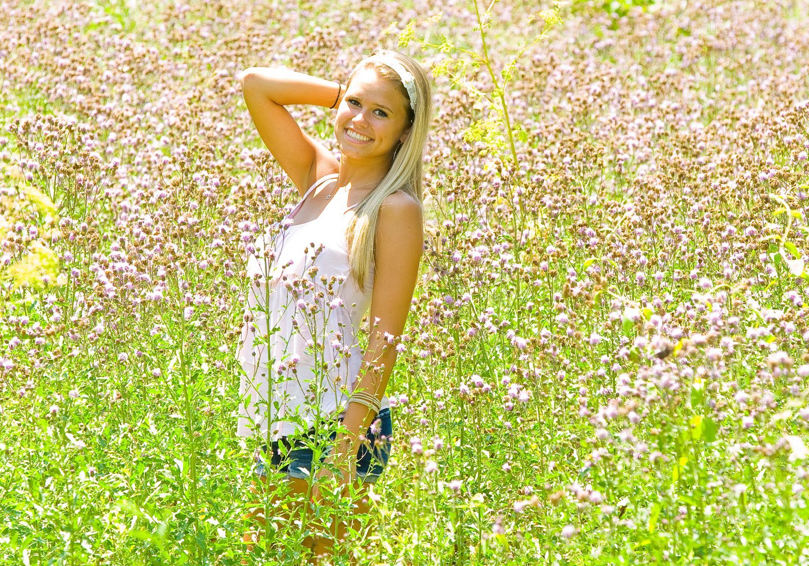Woman in a white top standing in a field of pink and white wildflowers with one hand behind her head.