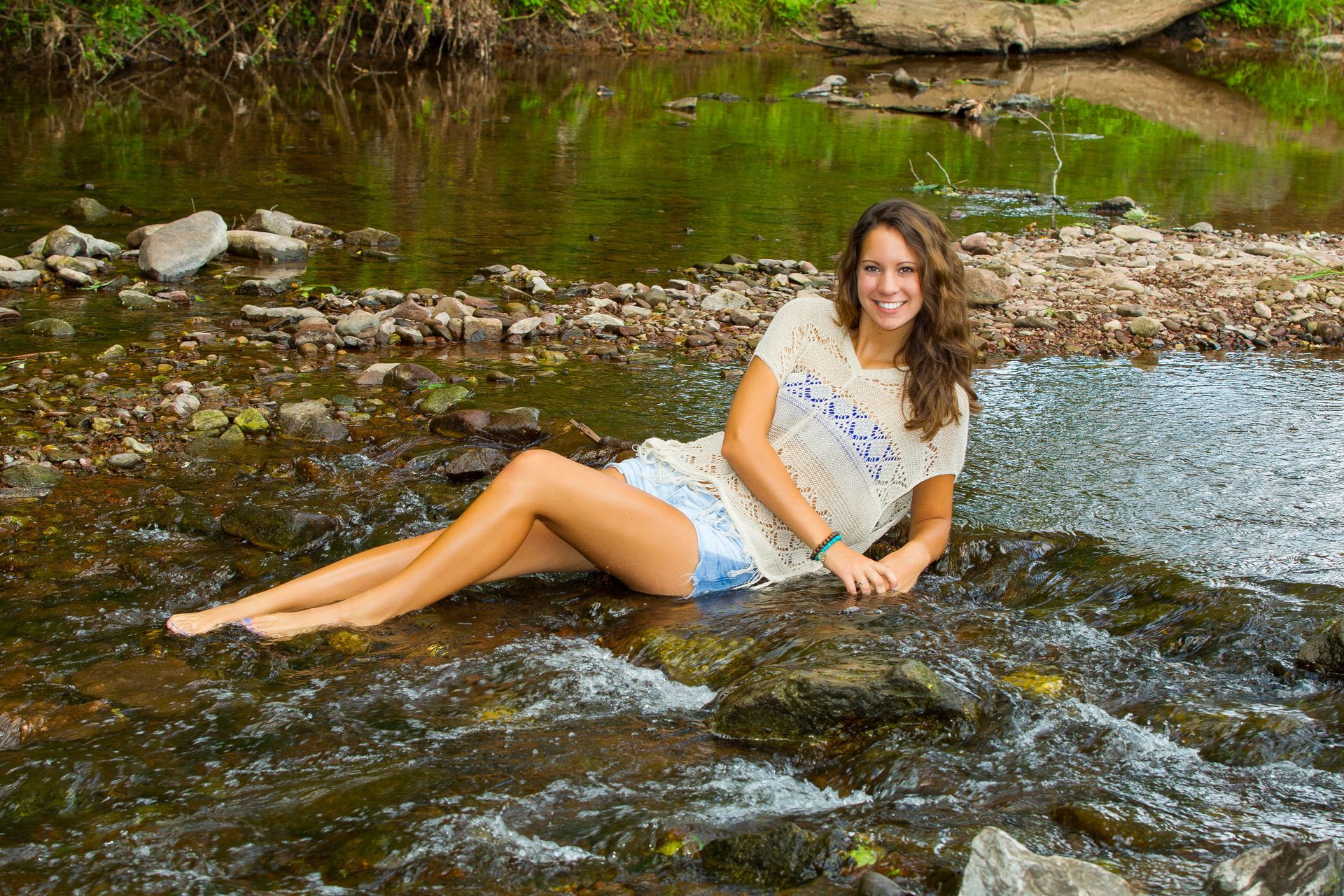 Woman reclining on rocks in a shallow stream, smiling in a white top and denim shorts.