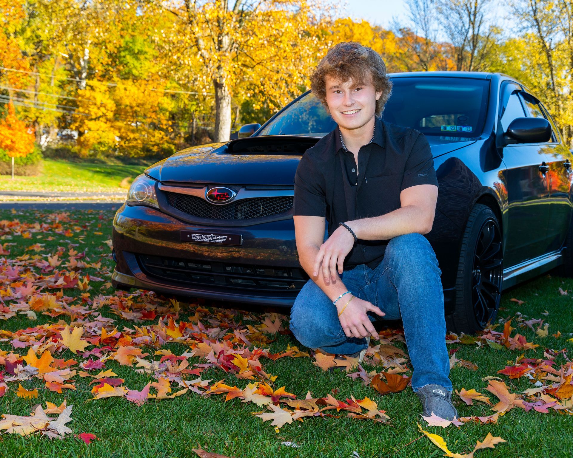 Person crouching beside a black car on a leaf-covered lawn in autumn.