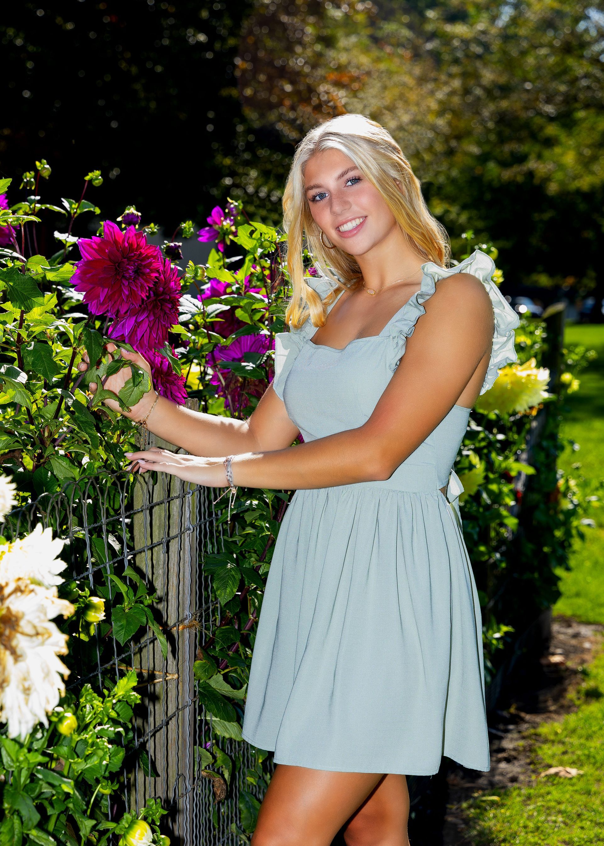Woman in a light blue dress standing by a garden railing, smiling beside bright pink flowers.