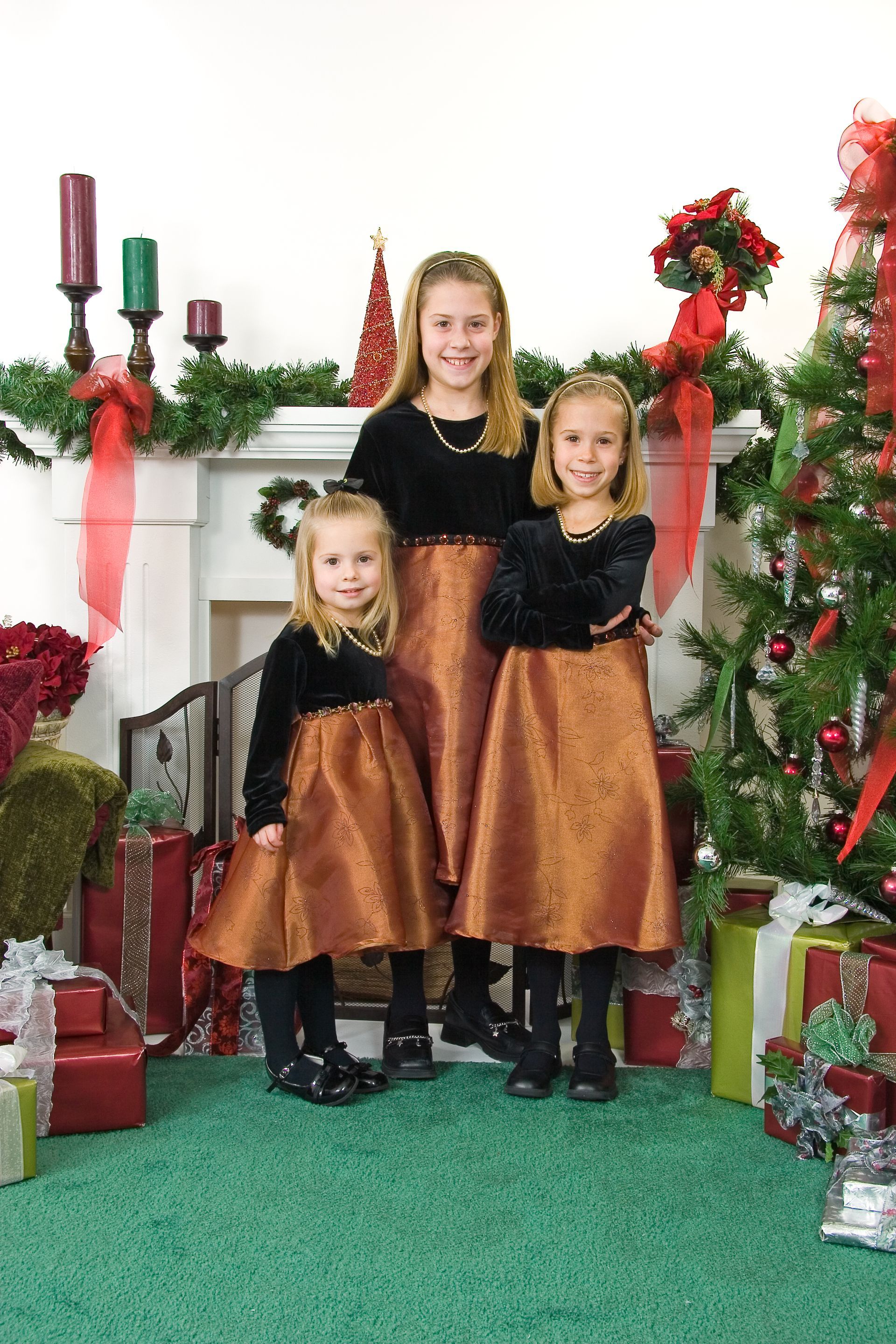 Three children in matching dresses posing by a decorated fireplace and Christmas tree