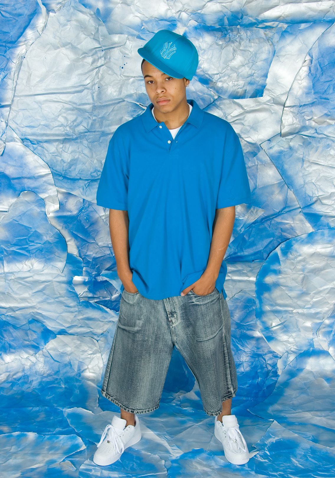 Man in blue polo and cap, gray shorts, and white sneakers standing against a blue textured backdrop