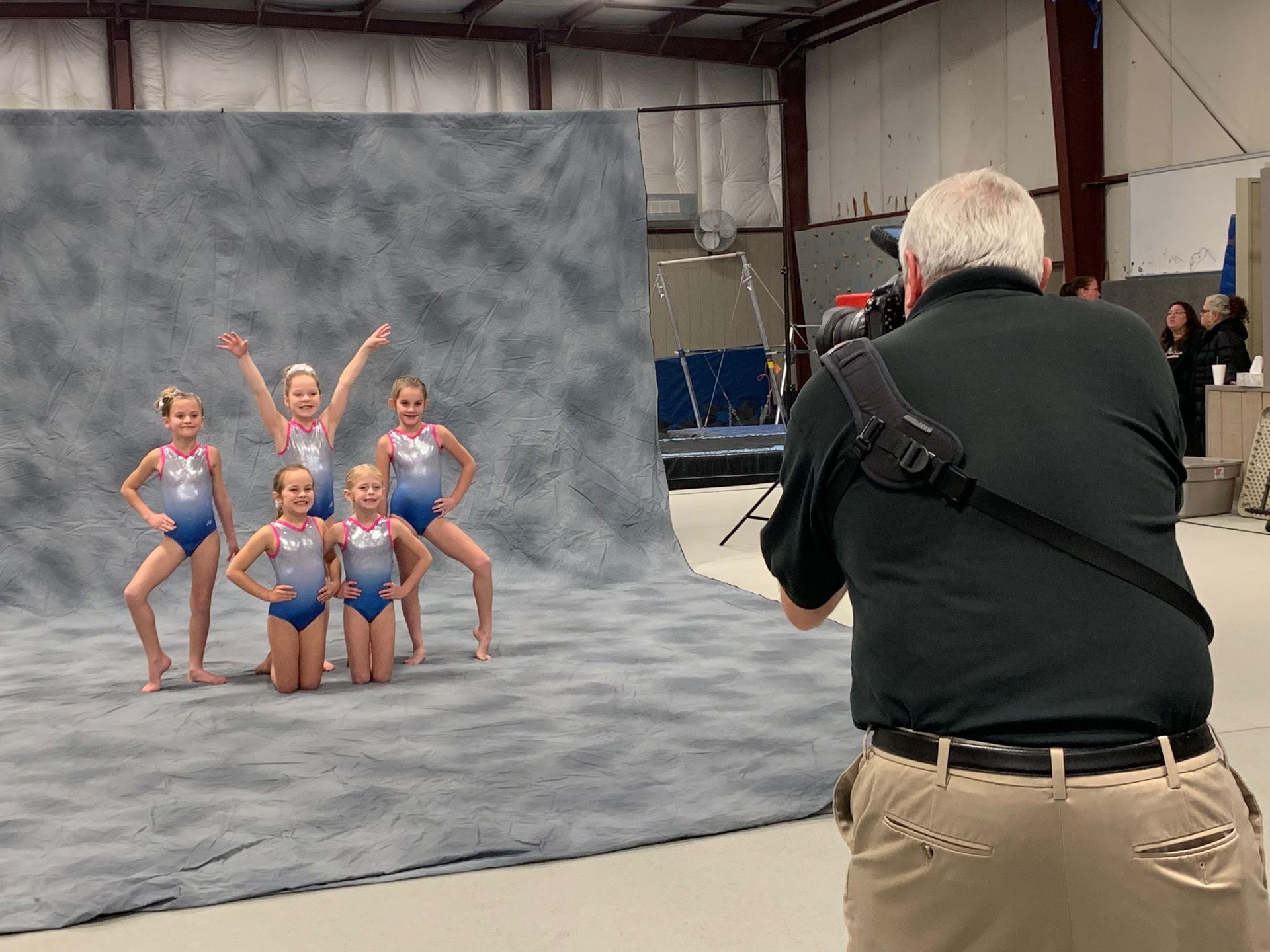 Photographer capturing six young gymnasts posing in blue leotards against a gray backdrop indoors