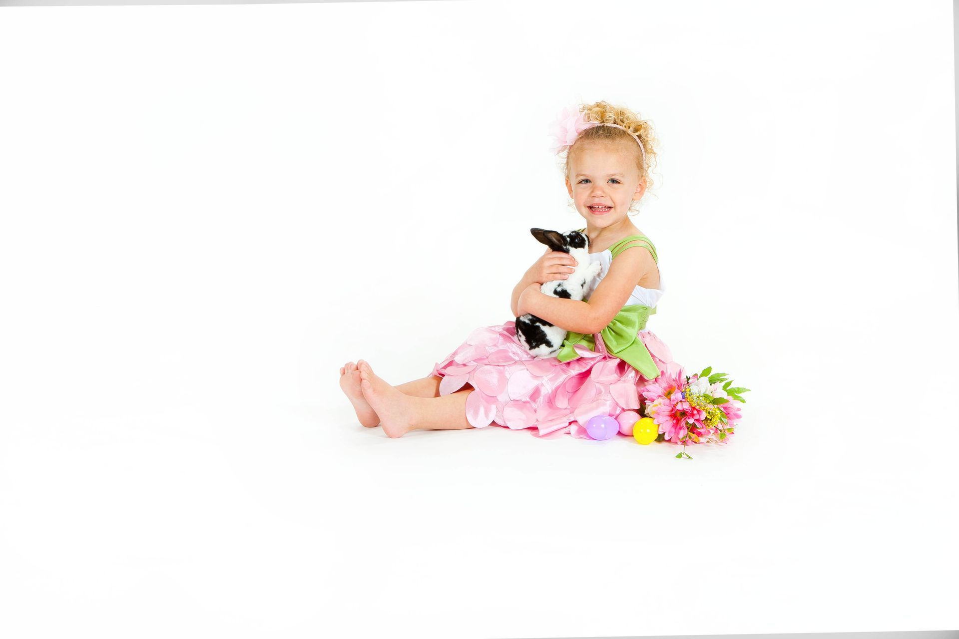 Young child in pink dress sitting on white background, holding a black-and-white cat with flowers beside her