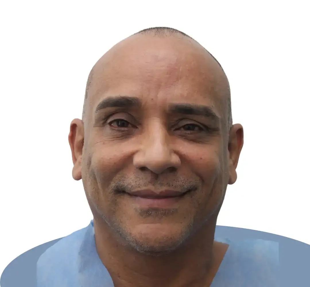 A man with a shaved head smiles, wearing a blue medical garment against a white background.