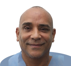 A man with a shaved head smiles, wearing a blue medical garment against a white background.