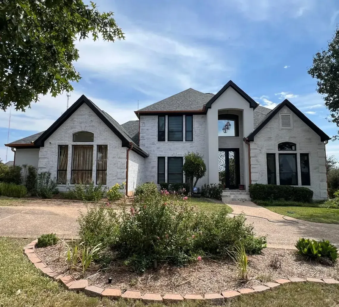 A two-story, white stone house with a multi-gabled roof, large windows, and a landscaped front garden under a blue sky.