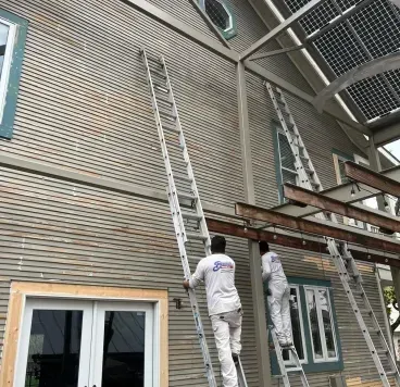 Two workers in white uniforms climb tall extension ladders against the light grey, textured exterior of a house.