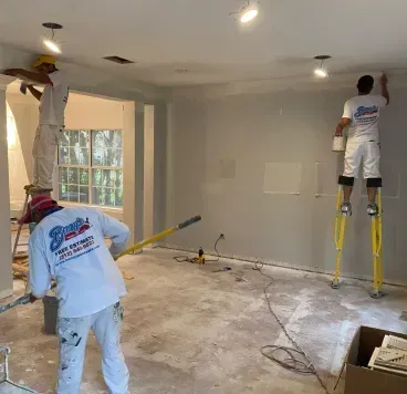 Three painters in matching white uniforms work on the walls and ceiling of a room under renovation.