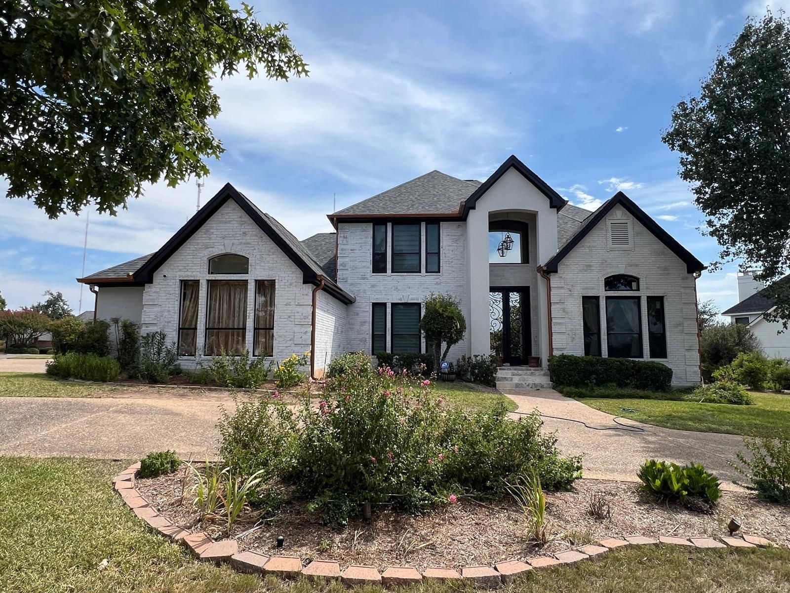 A two-story, white stone house with a multi-gabled roof, large windows, and a landscaped front garden under a blue sky.