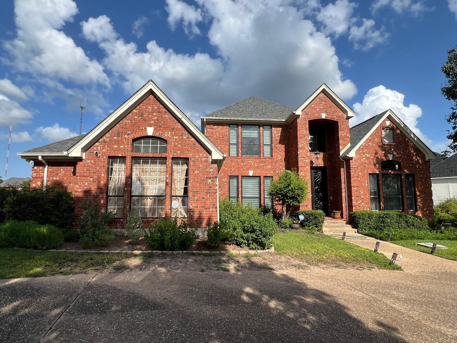A two-story red brick house with a gabled roof, dark-framed windows, and a front entryway under a blue sky with clouds.