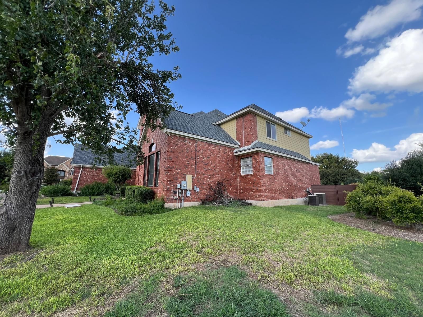 A two-story house with red brick and tan siding under a blue sky, viewed from a grassy yard with a large tree.