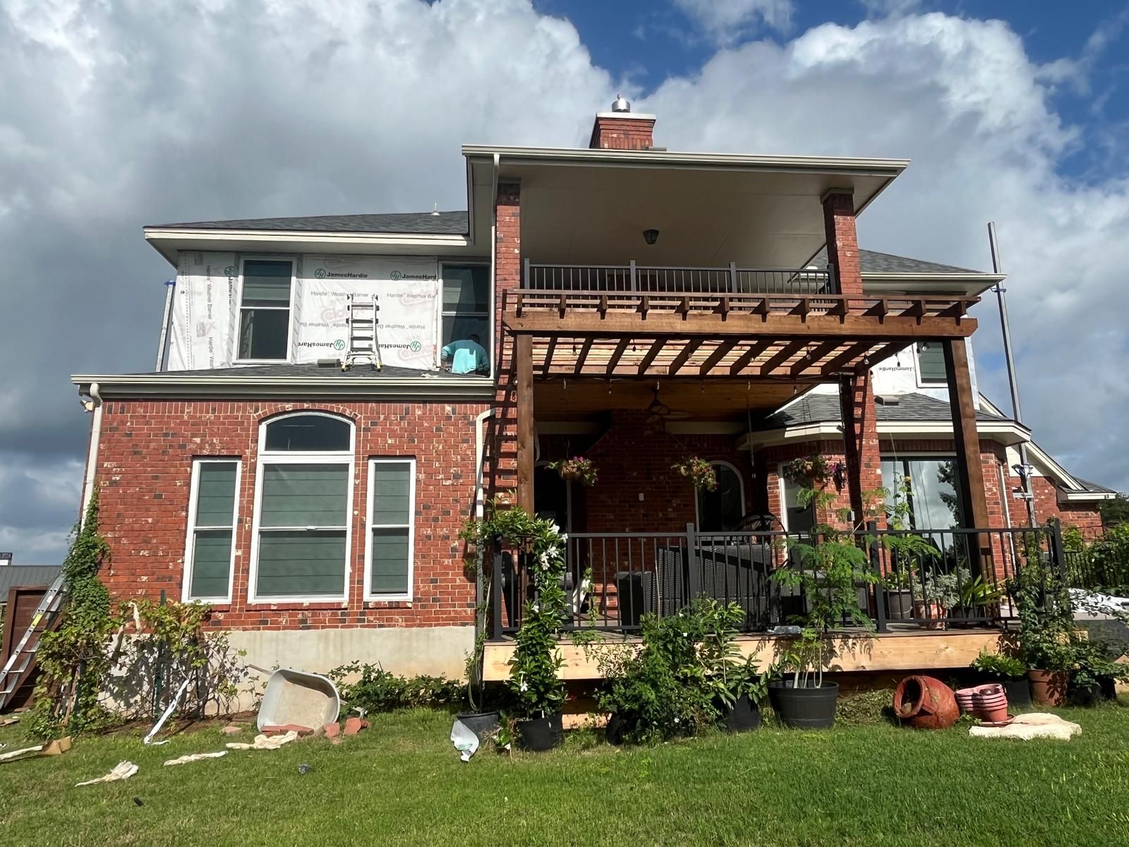 A two-story brick house with a partially completed upper level and a lower deck under a wooden pergola on a sunny day.