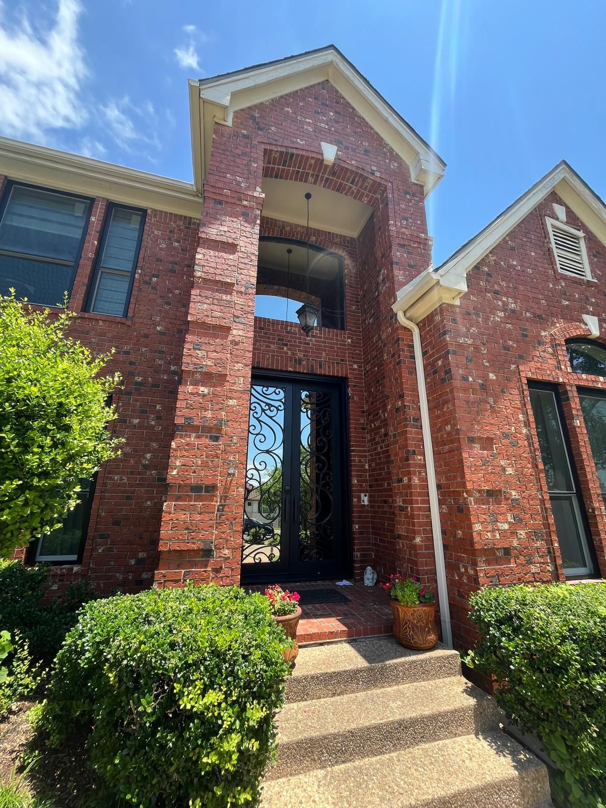 A brick house entrance featuring a tall, arched covered porch, iron-grille double doors, and stone steps under a blue sky.