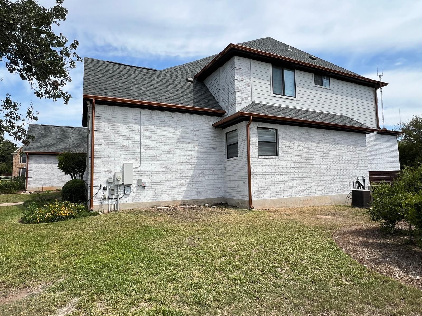 A two-story house with white brick exterior, dark grey roof, and copper gutters under a blue sky with light clouds.