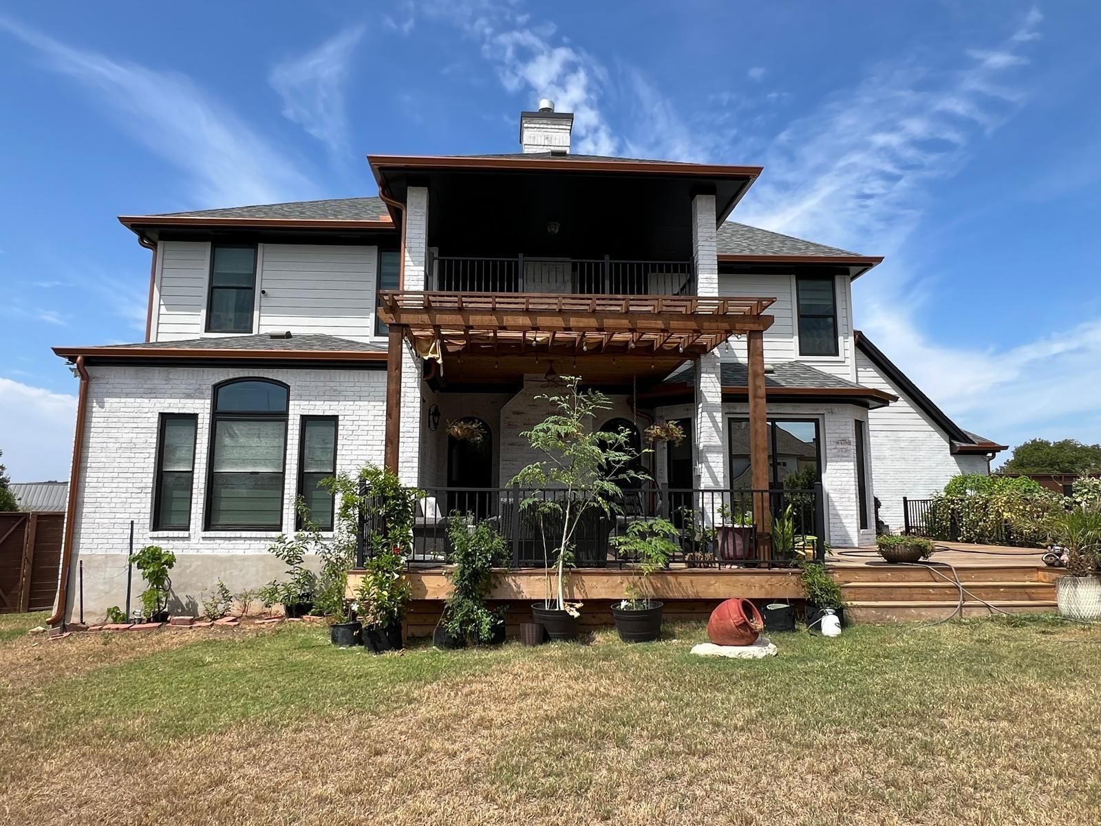 A two-story white brick house with a wooden pergola-covered back deck, surrounded by potted plants and a grassy lawn.