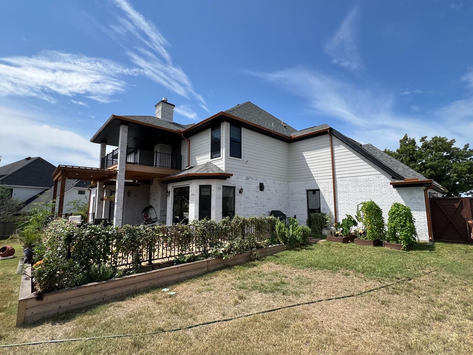 A two-story white brick house with a dark roof and a wooden back patio, set against a bright blue sky.