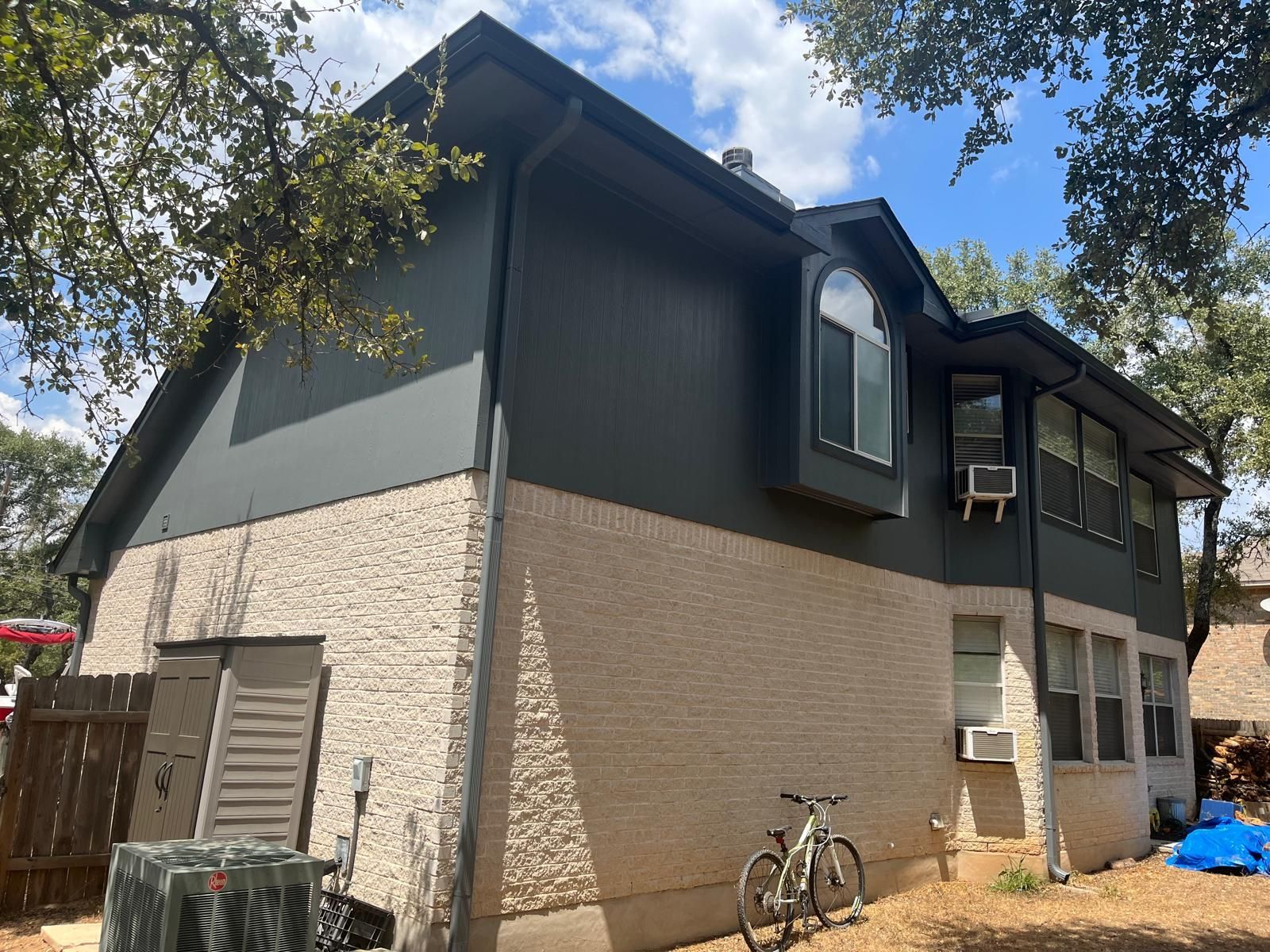 Two-story house with dark gray siding, light-colored brick, a bay window, and two air conditioning units in a yard.