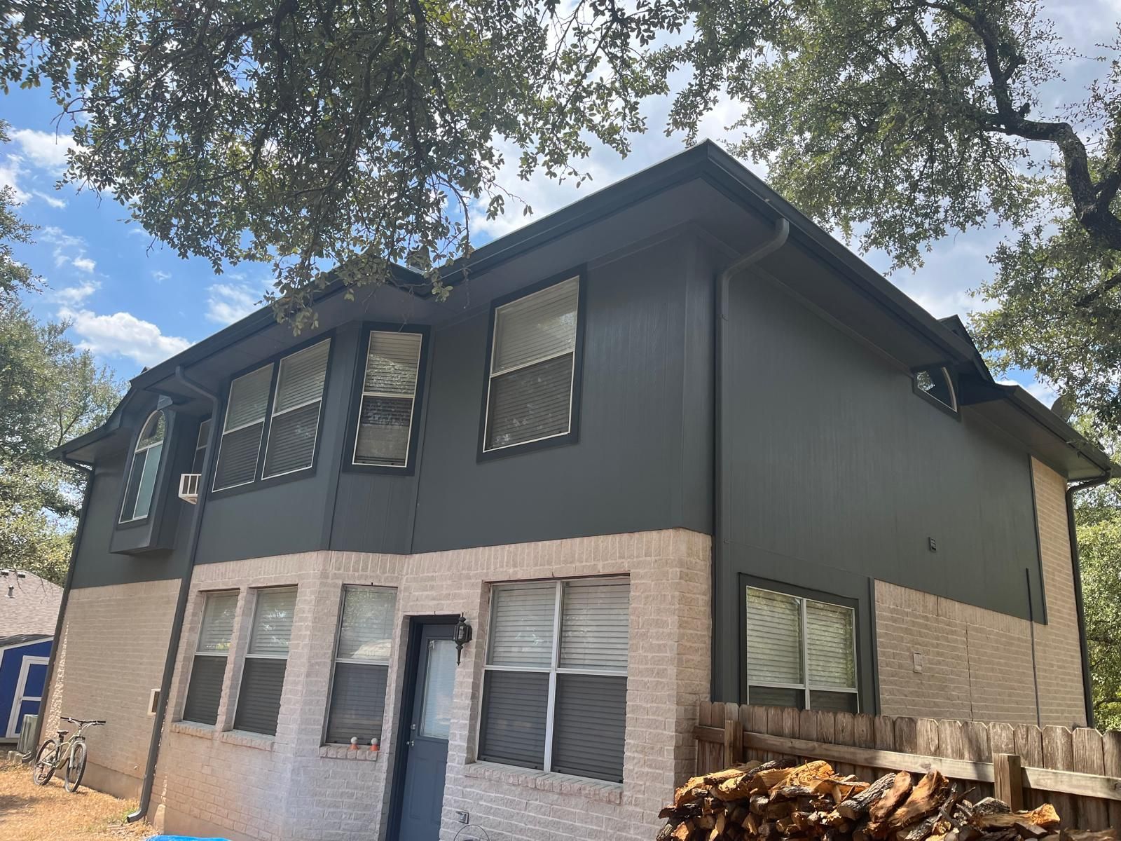 Two-story house with dark gray siding on the upper level and light-colored brick on the ground floor under leafy trees.