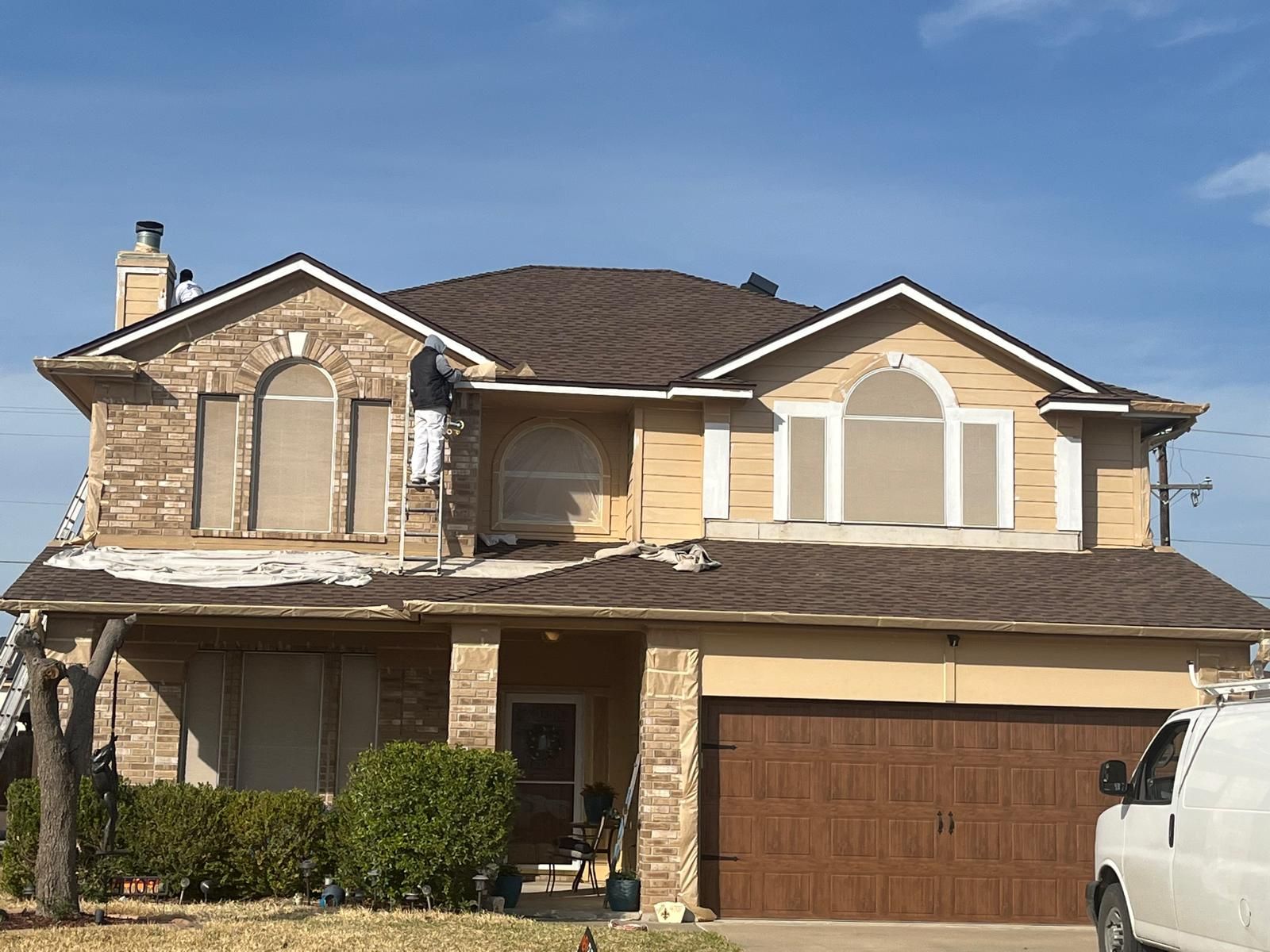 A two-story tan house with a brown roof and a brown garage, undergoing exterior repairs by a worker on a ladder.