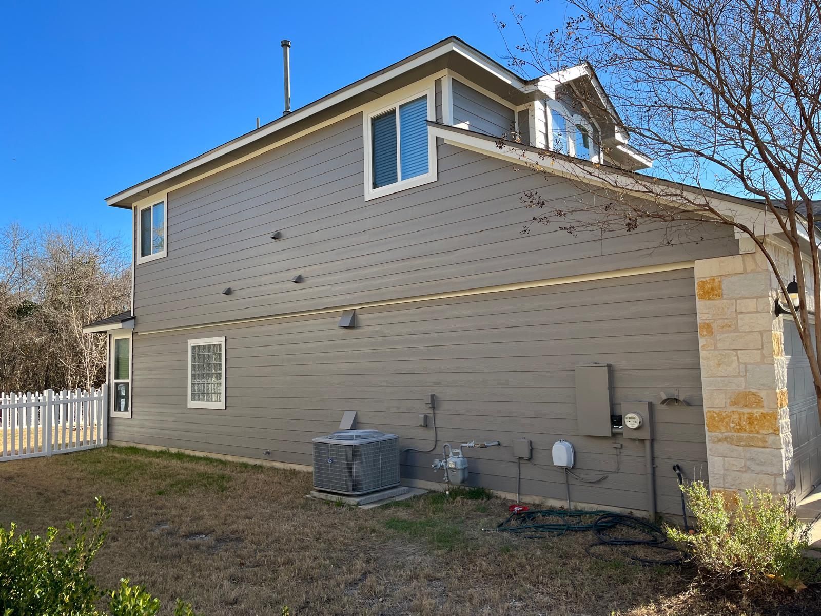 Side view of a two-story house with gray lap siding, stone accents, and an HVAC unit on the ground against a blue sky.