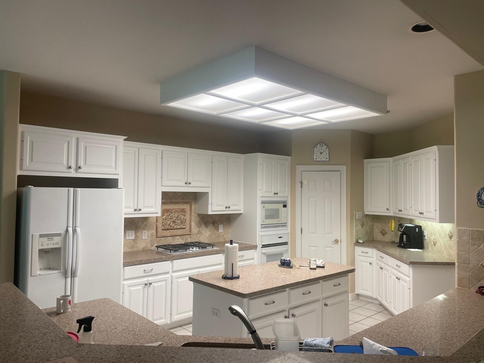 A spacious kitchen with white cabinets, beige countertops, a central island, and a large recessed light fixture.