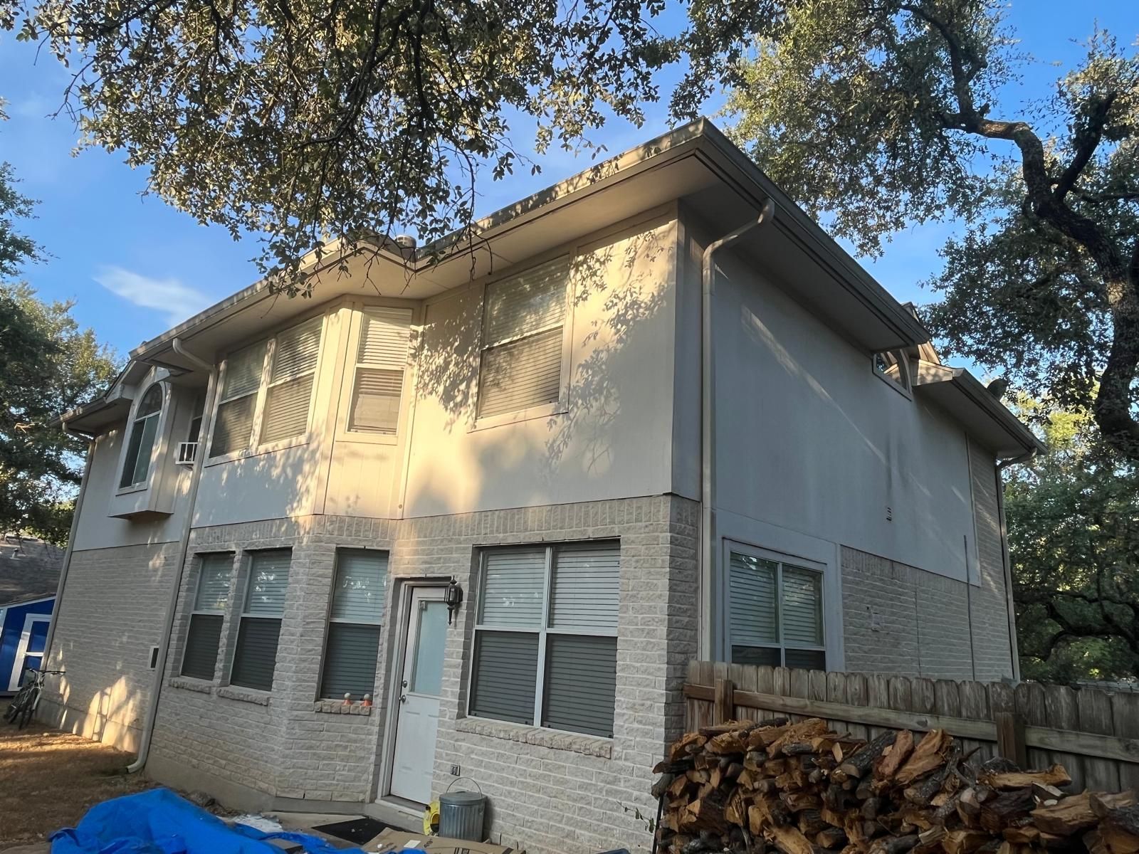 A two-story house with a white brick lower level, light-colored upper siding, and a wooden fence in the foreground.