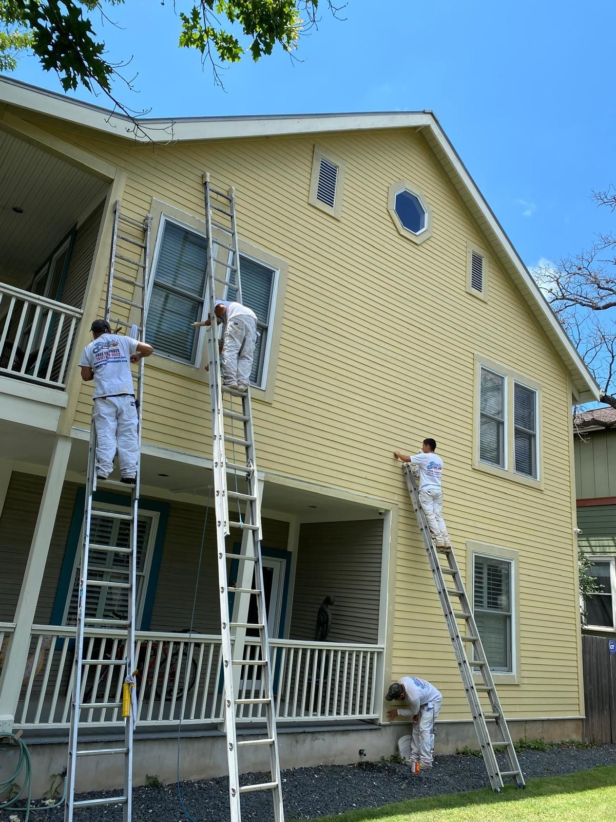 Four painters in white work uniforms use ladders to paint the exterior of a two-story yellow house.