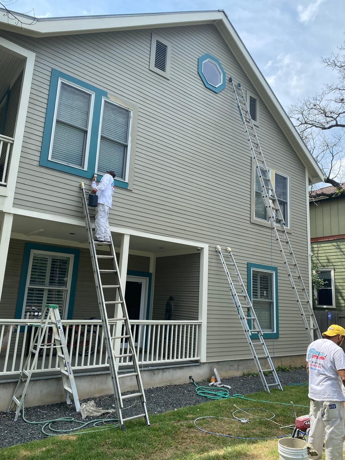 Workers in white coveralls use ladders to paint the exterior trim of a two-story gray house.