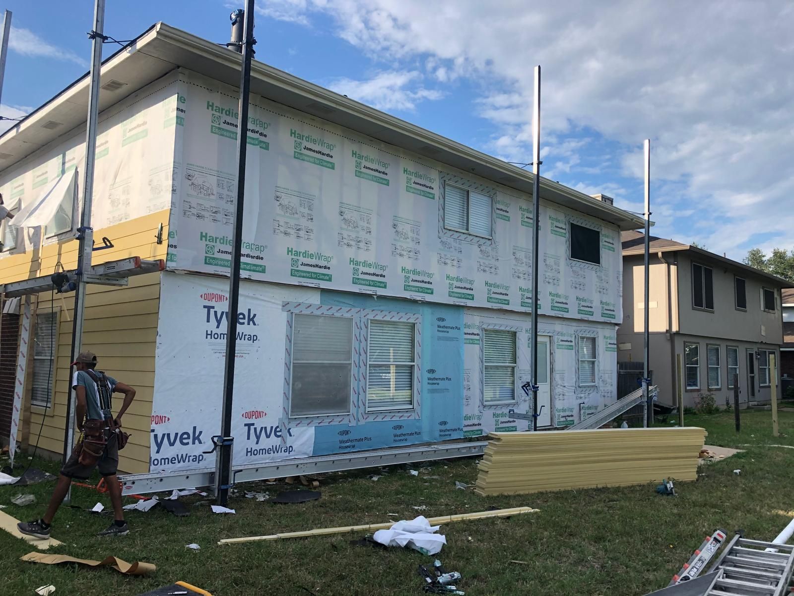 A worker stands near a two-story residential building undergoing exterior renovation with Tyvek house wrap installed.