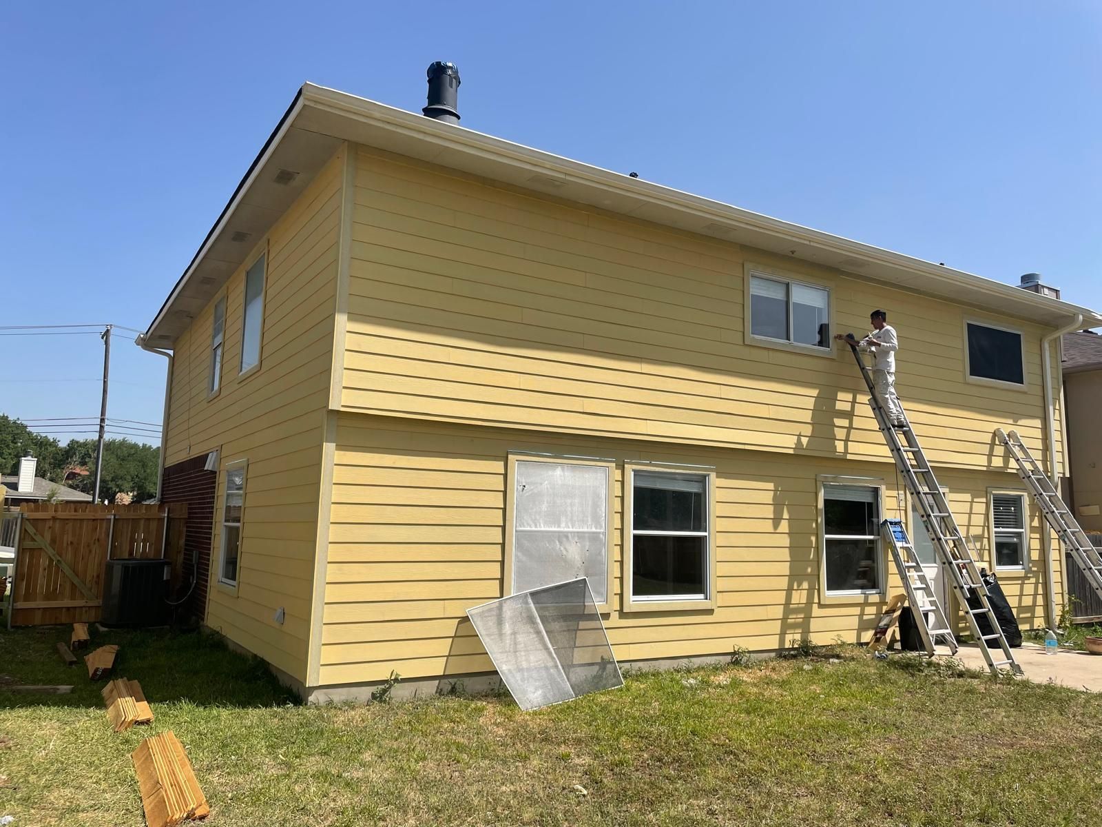 A person in protective gear on a ladder paints the yellow siding of a two-story house under a clear blue sky.
