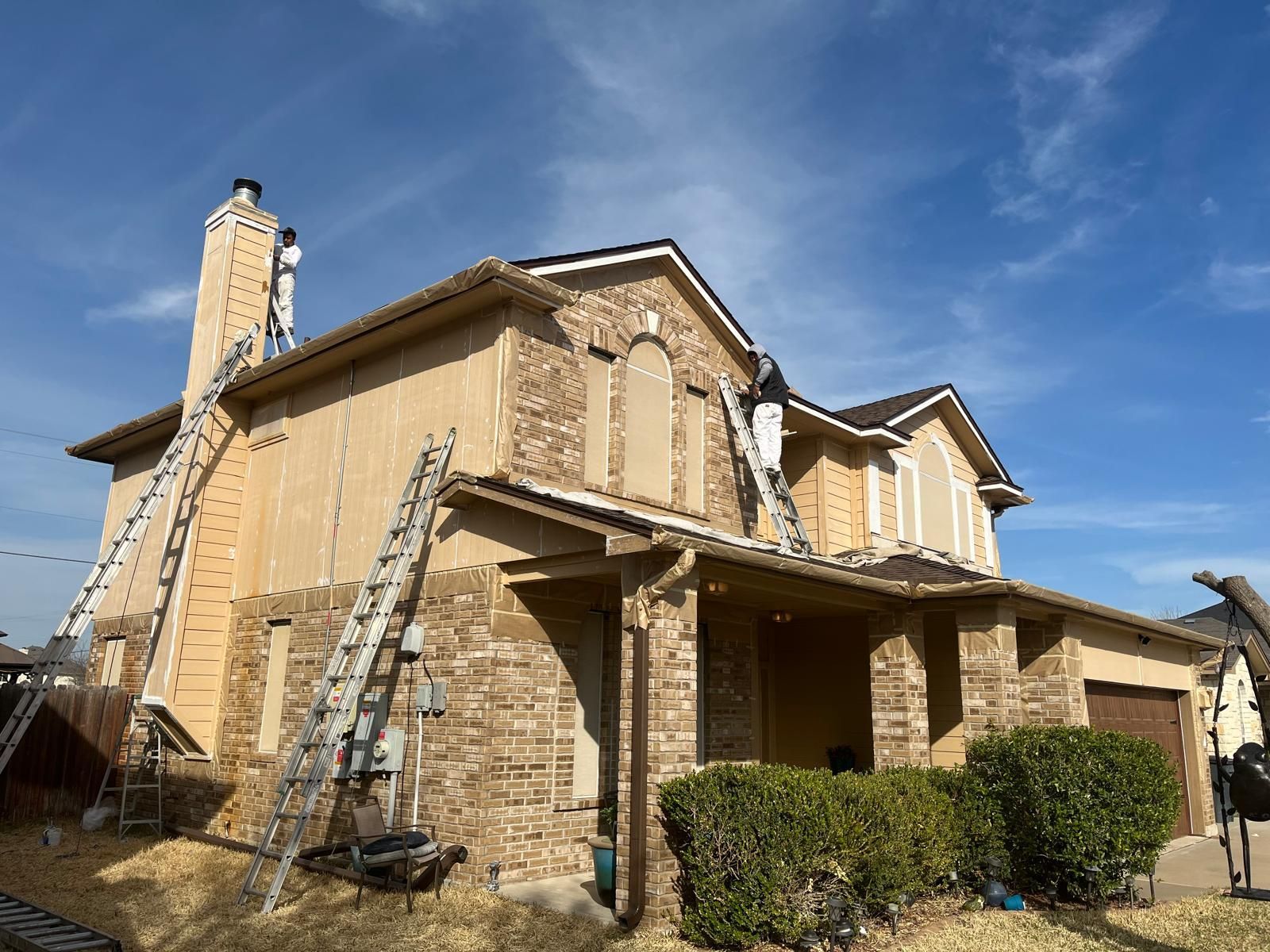 Two workers in protective suits use ladders to work on the exterior siding of a two-story home with stone and stucco.