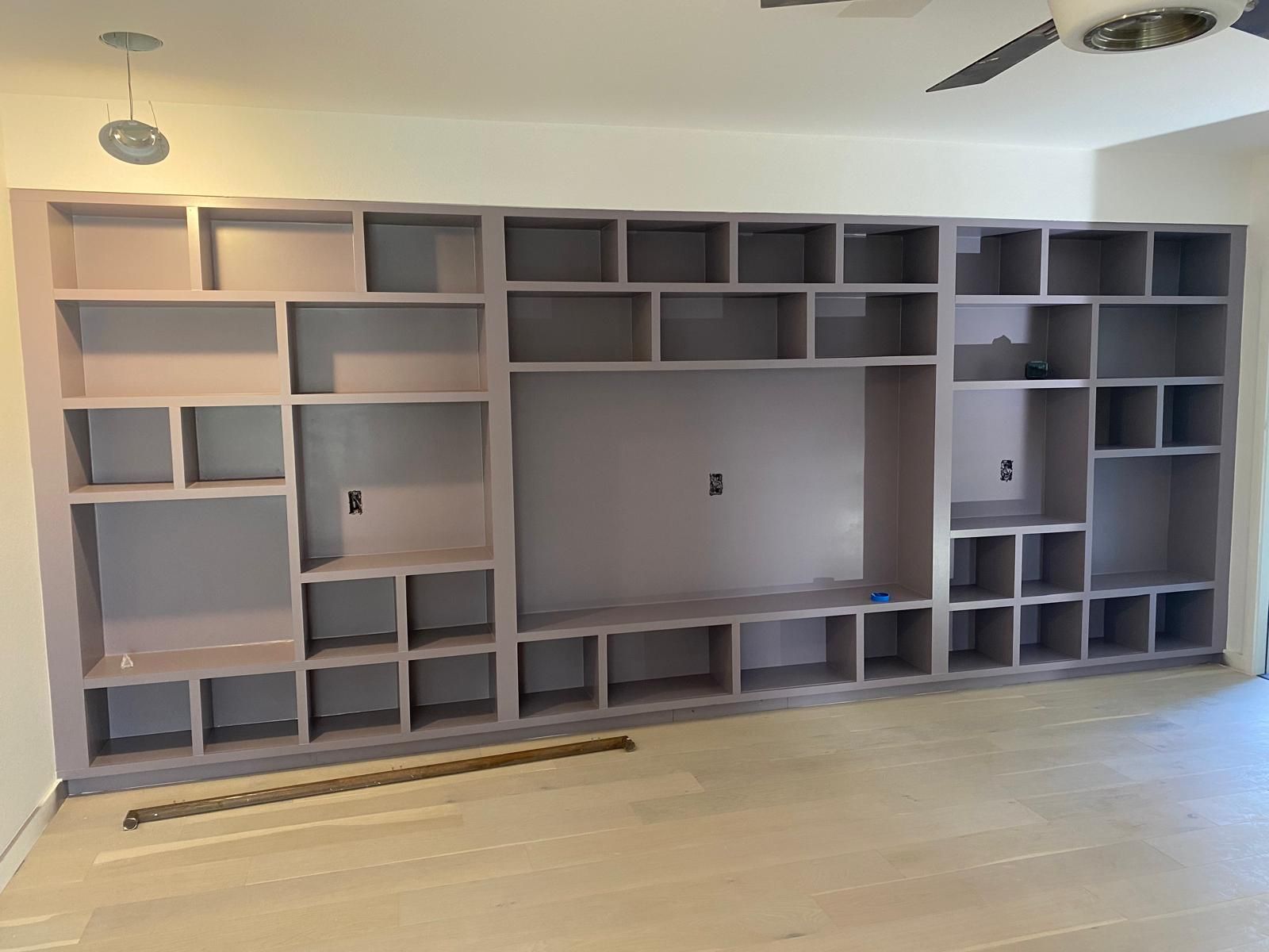 A large, built-in wall unit with many shelves and a central TV niche, painted in a light purple color in a room.